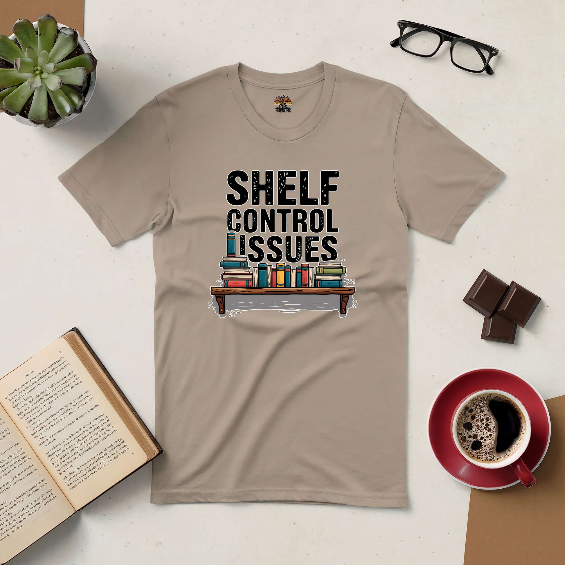 Shelf Control Issues tee with books graphic, surrounded by coffee, chocolate, and open book on a table.