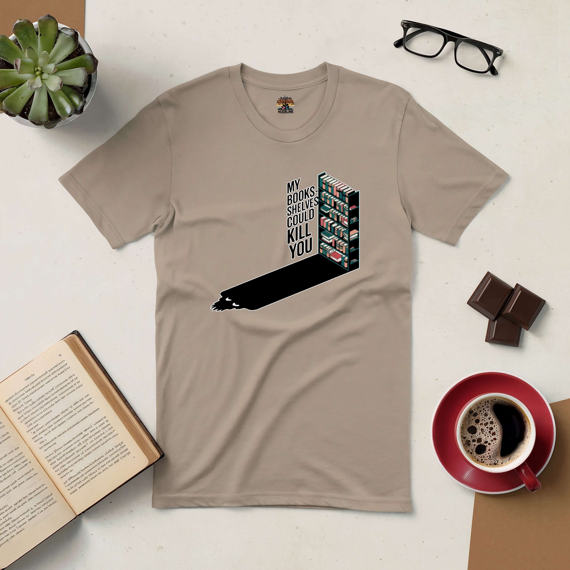 Tan t-shirt with "My Bookshelves Could Kill You" design, surrounded by books, coffee, glasses, and a plant, for book lovers.