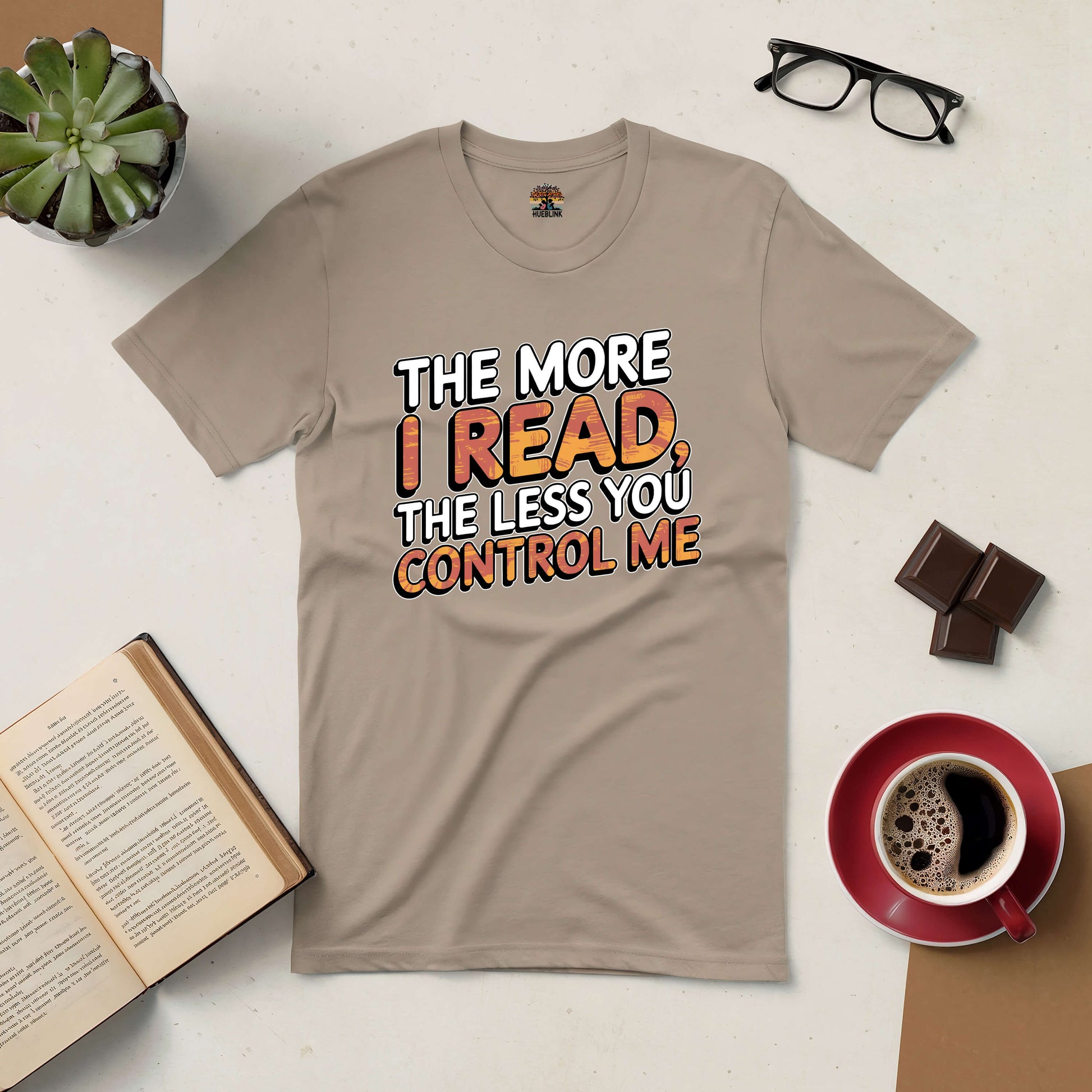 Beige graphic tee with "The More I Read, The Less You Control Me" text, surrounded by coffee, glasses, and a book.