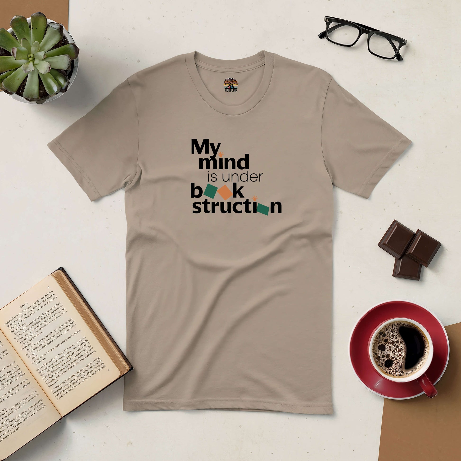 Beige tee with "My Mind is Under Book-struction" text design, celebrating reading and continual knowledge growth, surrounded by coffee and books.