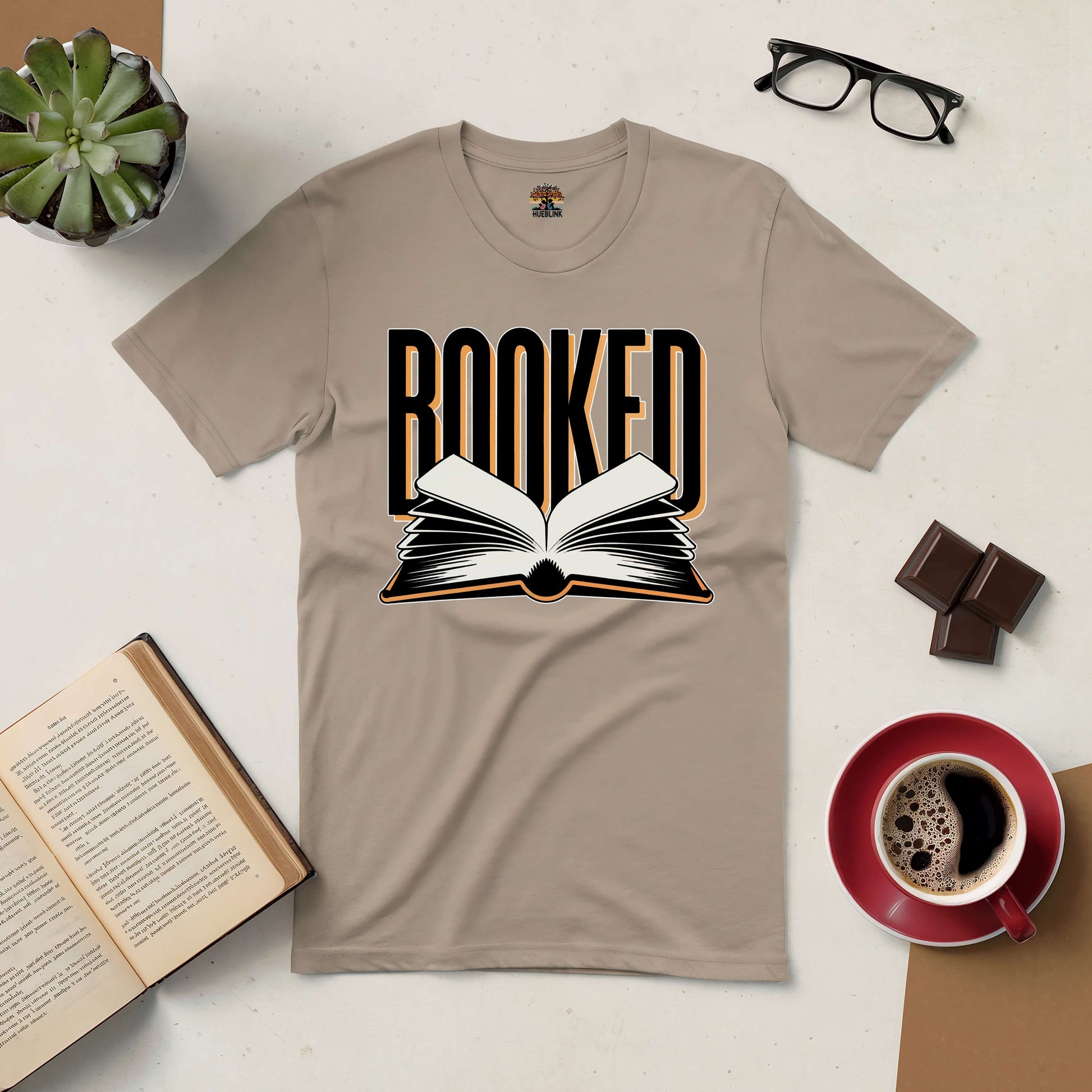 Beige "Booked" tee with open book design, surrounded by coffee, chocolates, glasses, an open book, and a plant. Perfect for book lovers.