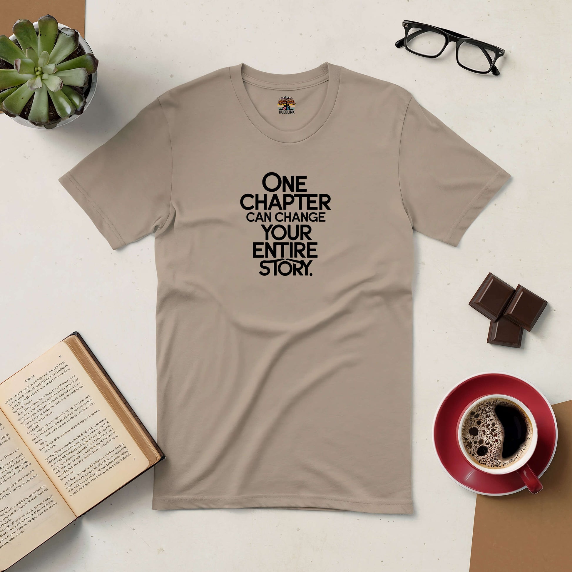 Inspirational tee with "One Chapter Can Change Your Entire Story" text, shown with coffee, book, glasses, plant, and chocolate on table.