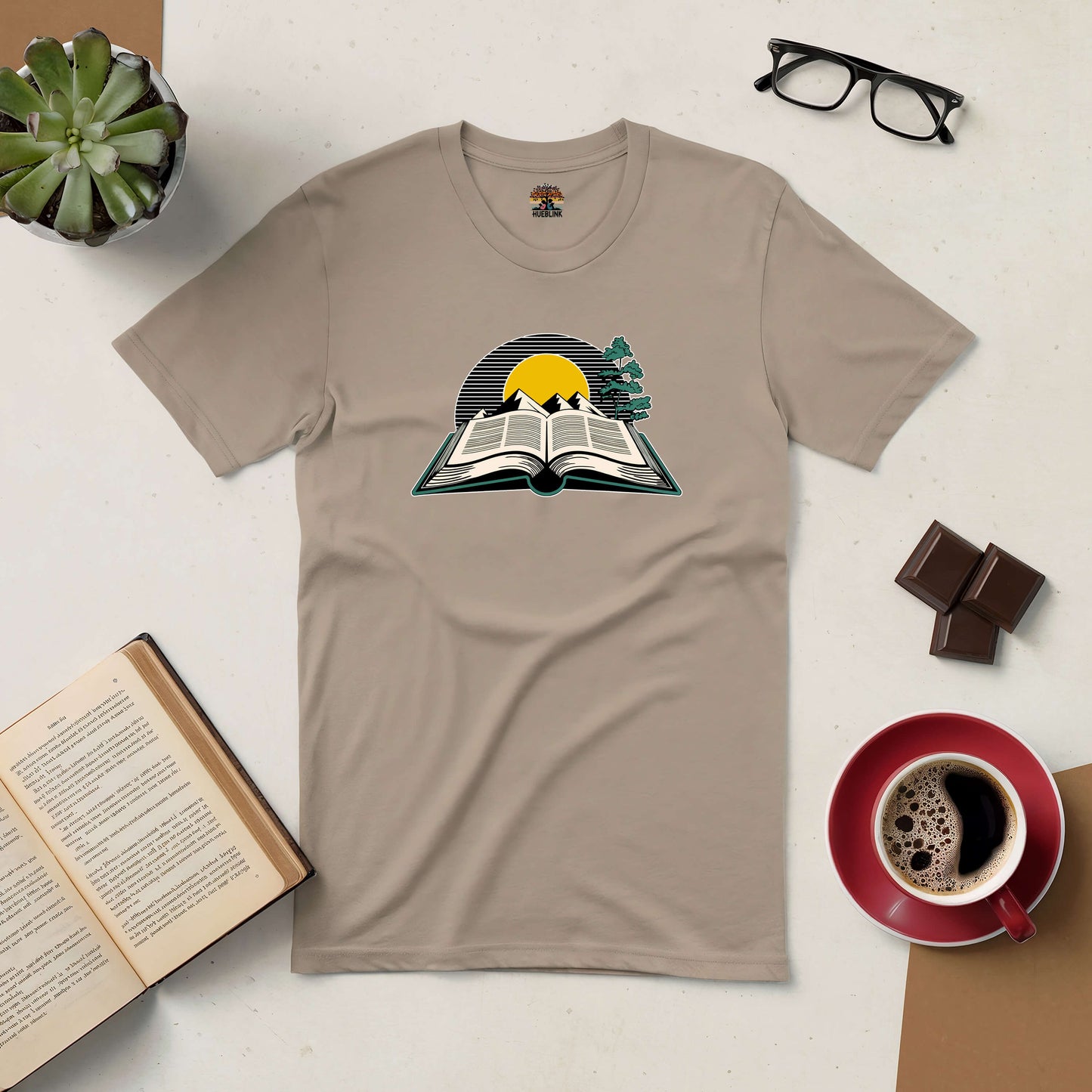 Beige tee featuring a sunrise over an open book, symbolizing new adventures. Displayed on a table with glasses, a book, coffee, and chocolates.