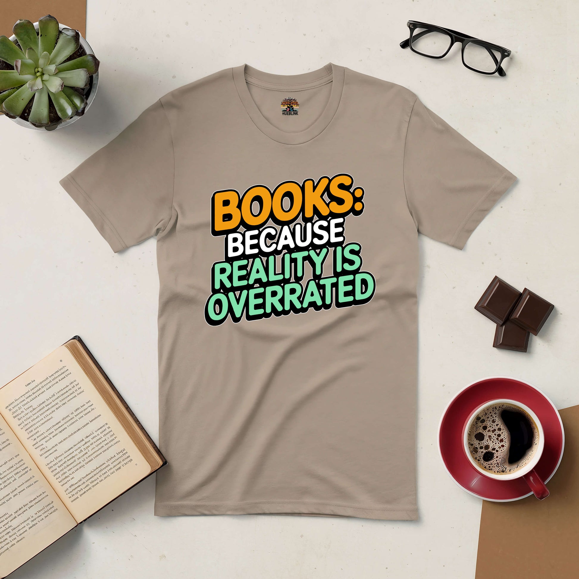 Beige t-shirt with "Books: Because Reality Is Overrated" text, surrounded by a book, coffee, glasses, and plant.