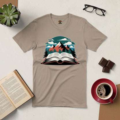 Beige tee with mountain and open book design, surrounded by book, coffee, glasses, plant, and chocolate on a table.