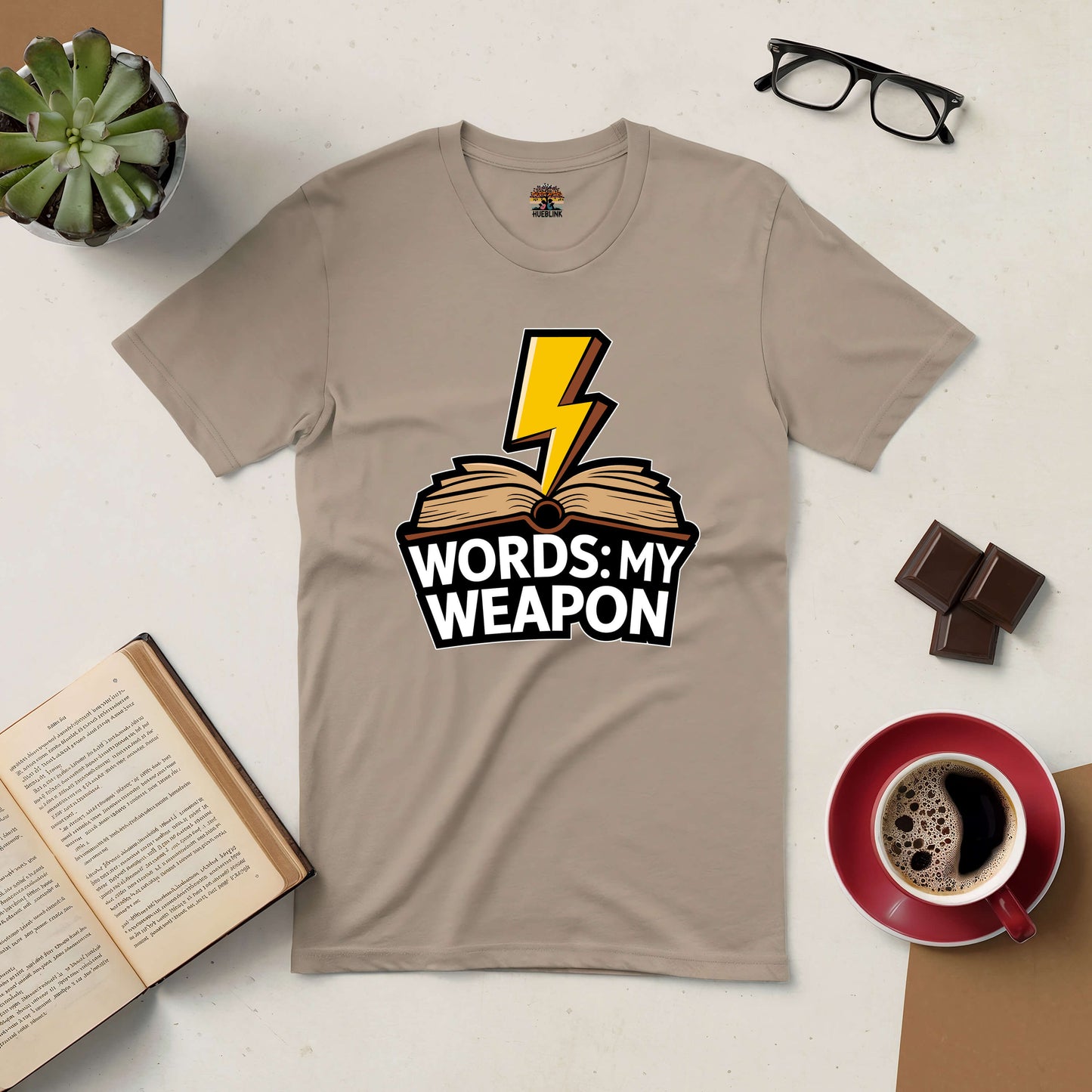 Tan Words: My Weapon Tee featuring a lightning bolt and book design, emphasizing the power of language over force, displayed on a table.