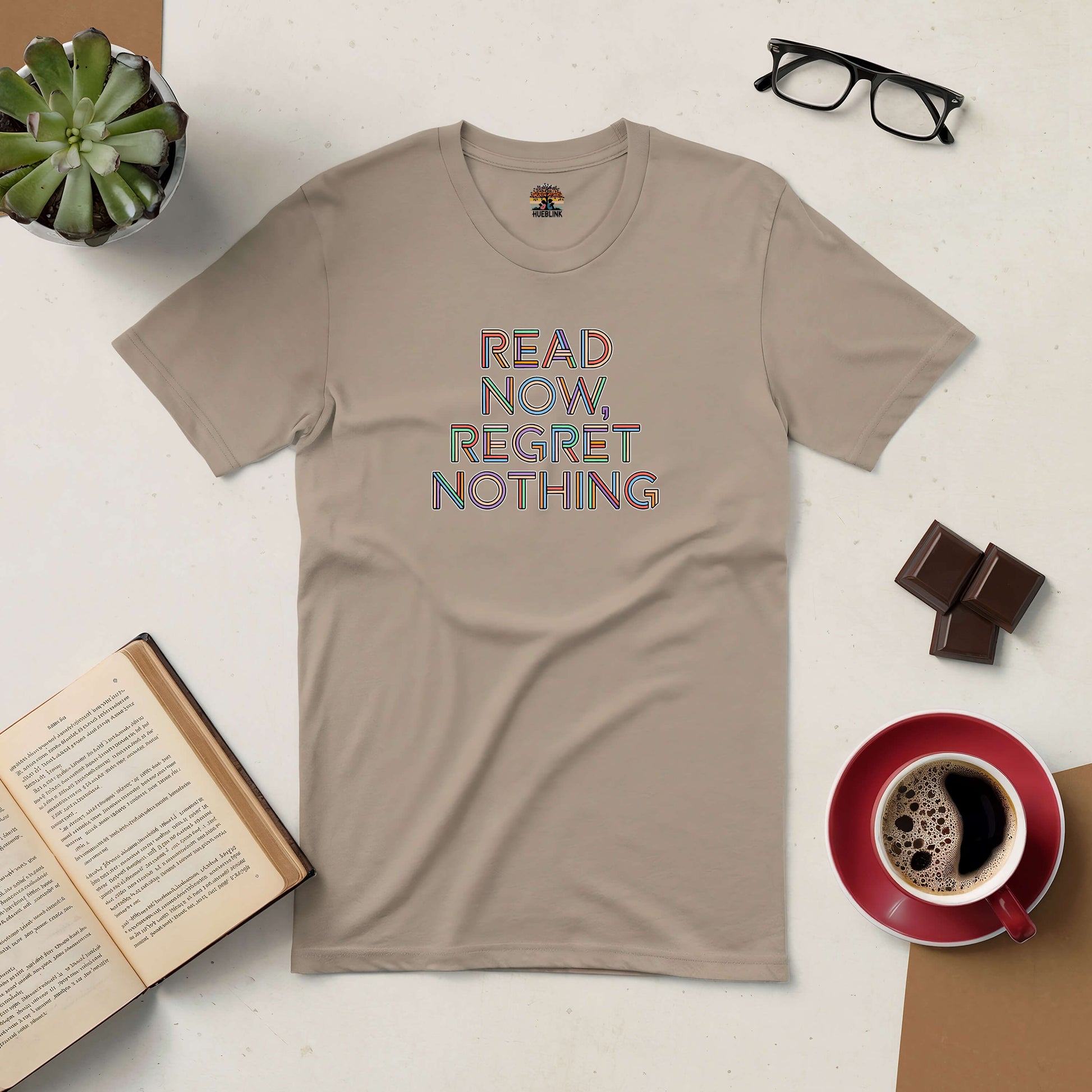 Beige "Read Now, Regret Nothing" t-shirt with coffee, chocolate, glasses, and an open book on a table, book lover's delight.