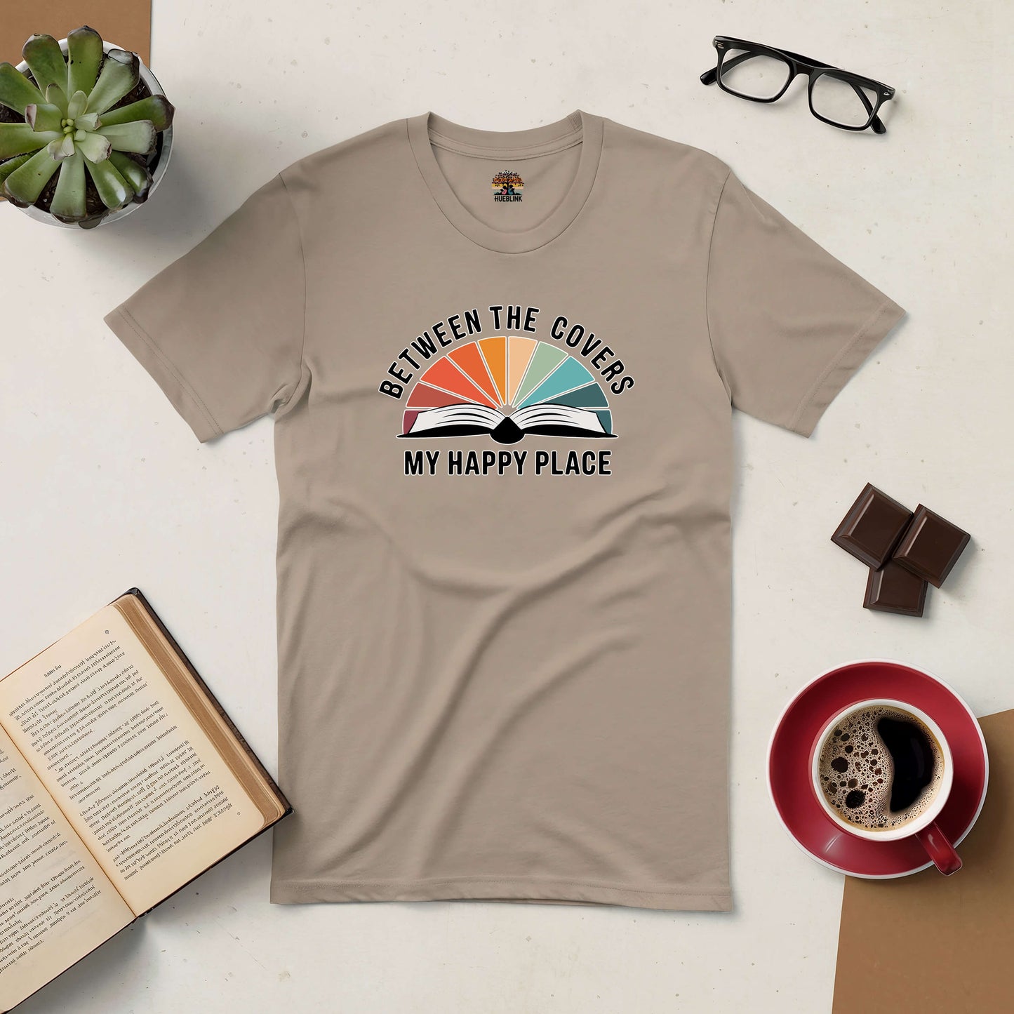 Beige "Between the Covers: My Happy Place" t-shirt with open book graphic, surrounded by coffee, chocolate, glasses, open book, and plant.