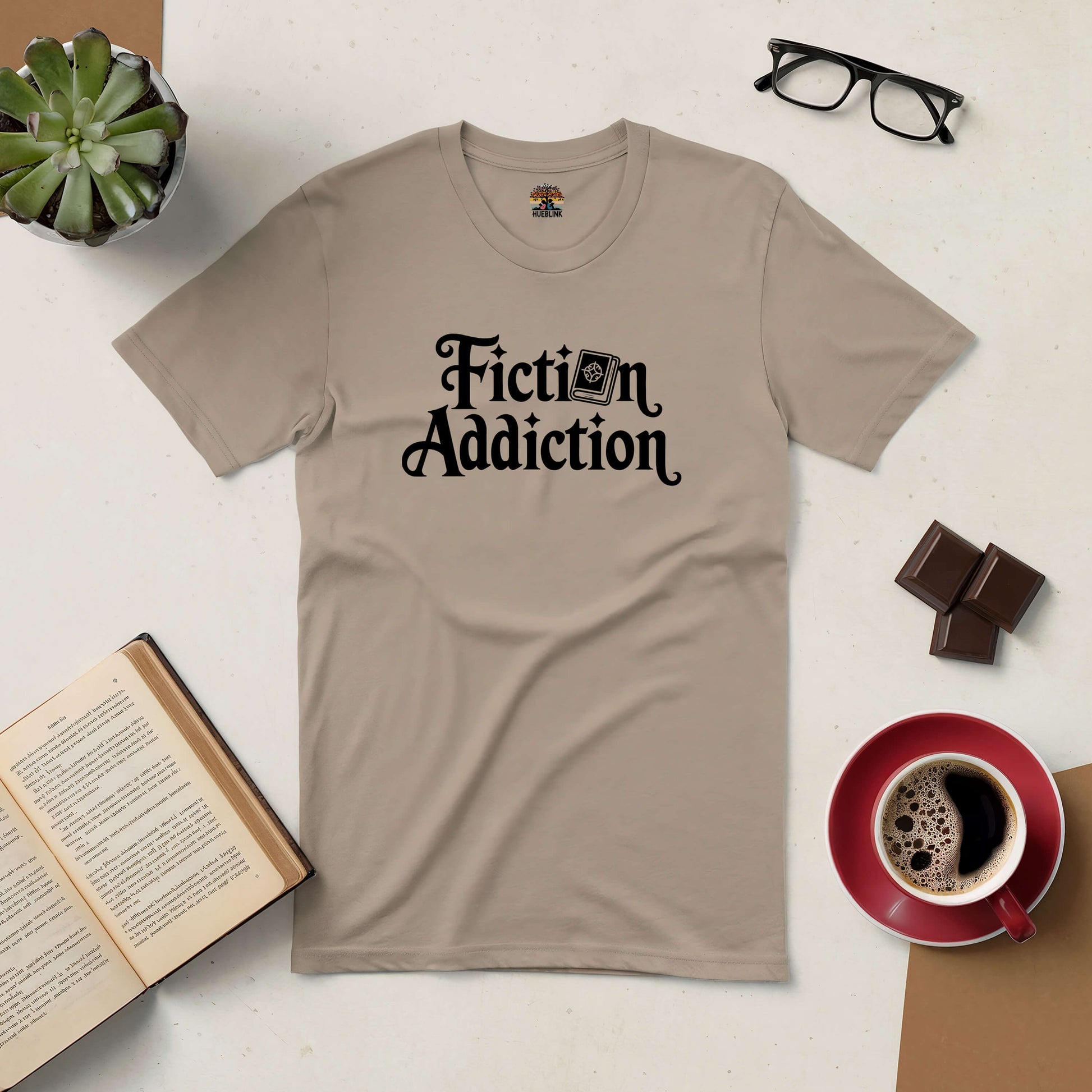 Fiction Addiction Tee with book design, surrounded by reading essentials: open book, coffee, chocolate, glasses, and plant.