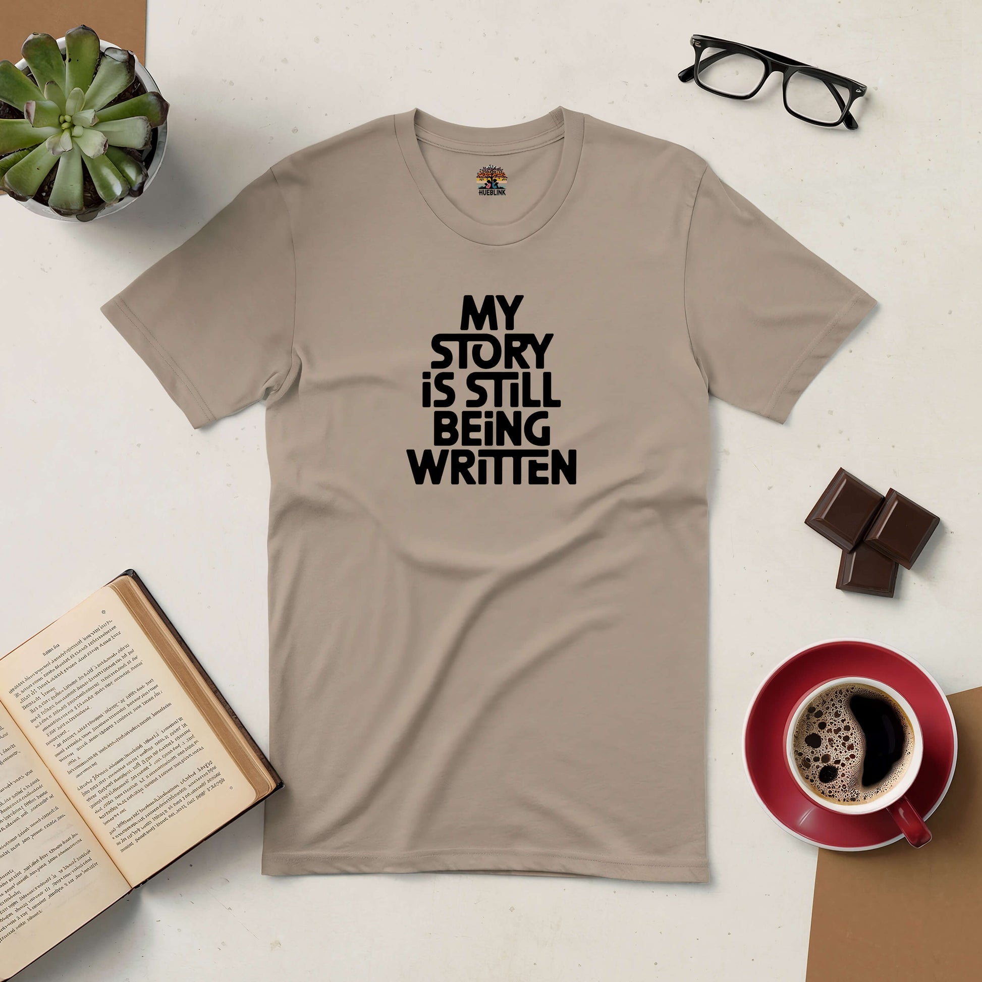 Beige "My Story Is Still Being Written" tee on table with coffee, chocolate, book, glasses, and succulent plant.