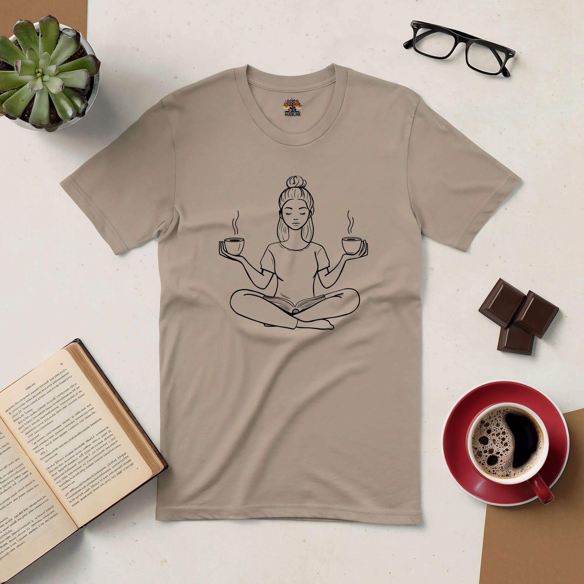 Beige tee featuring a zen design with a person meditating, holding coffee and book, surrounded by coffee cup, book, and glasses.