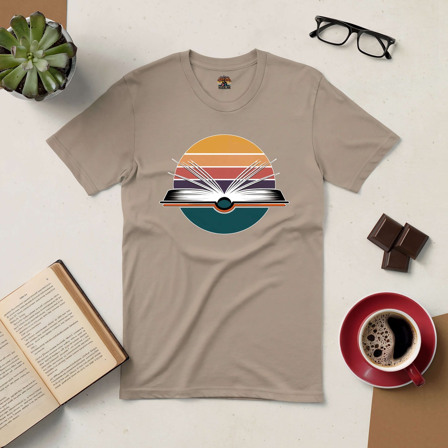 Beige tee with artistic book design, symbolizing endless possibilities. Coffee, glasses, book, and plant in the background.