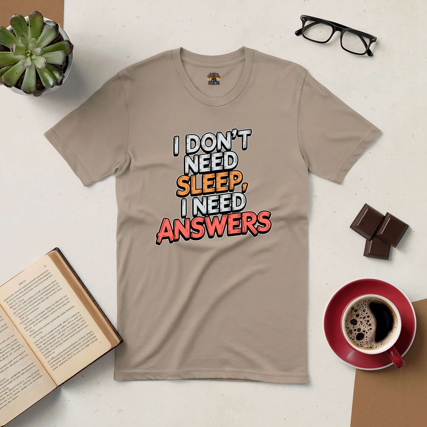 Beige tee with "I Don't Need Sleep, I Need Answers" slogan amidst books and coffee, perfect for curious night owls.