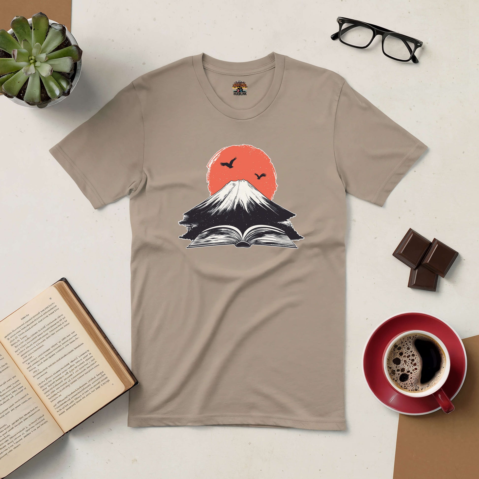 "Eruption of Imagination Tee featuring a volcano and book design, surrounded by coffee and open books, symbolizing adventure and creativity"