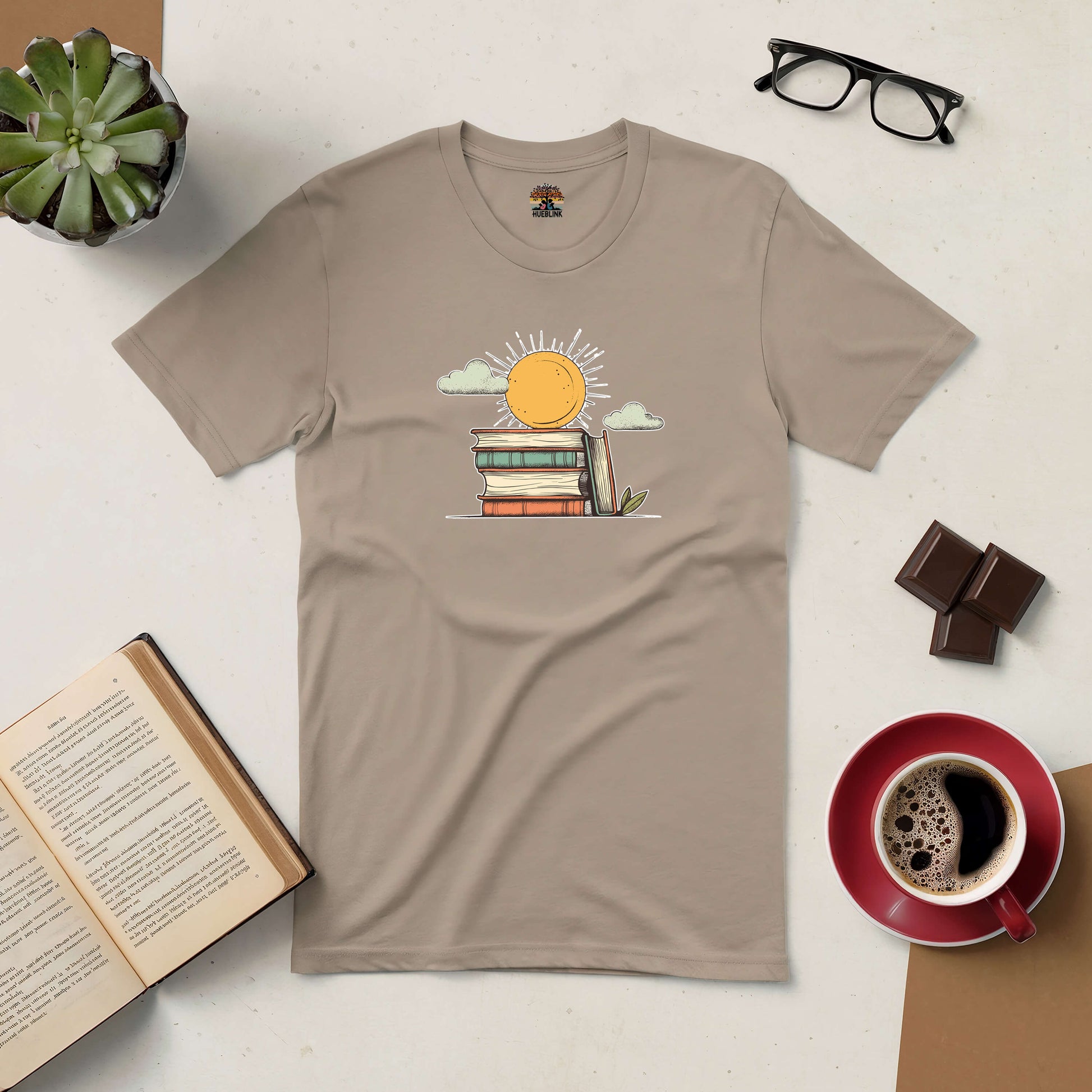 Beige Literary Sunshine tee with a sun and book design, surrounded by coffee, glasses, plant, and open book on a table.