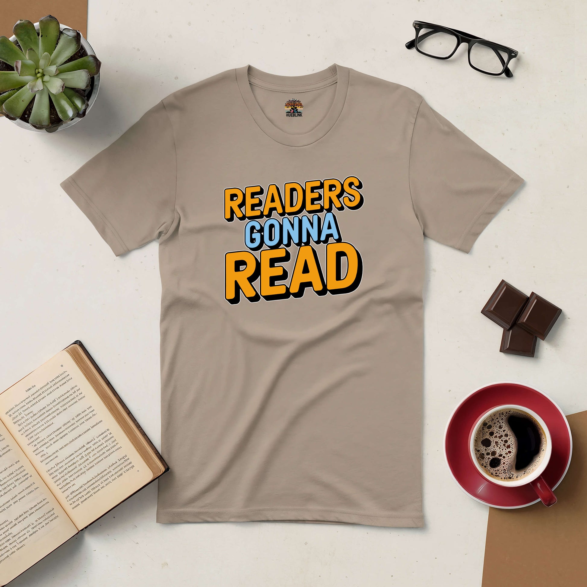 Beige "Readers Gonna Read" tee on a table with coffee, chocolate, glasses, plant, and an open book, perfect for book lovers.