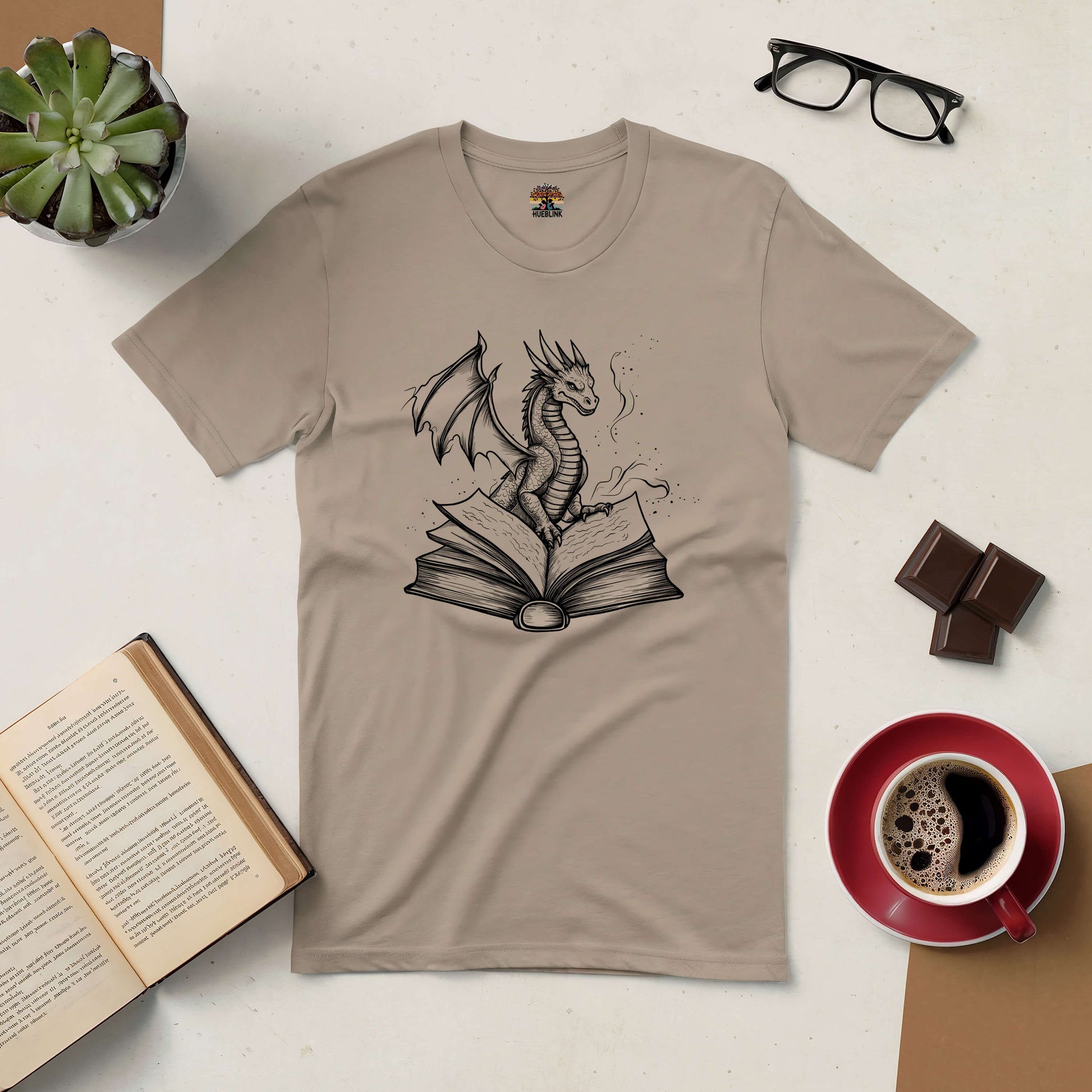 Beige Literary Dragon Tee with a dragon emerging from an open book, surrounded by coffee, chocolate, and reading items.