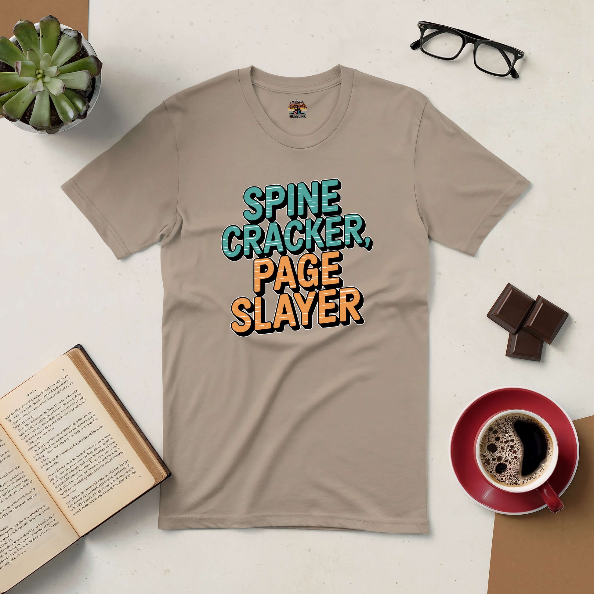 Tan "Spine Cracker, Page Slayer" tee for avid book lovers with coffee, glasses, and open book on a table.