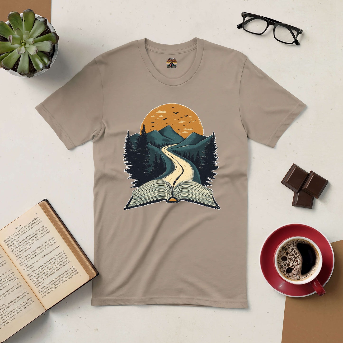 Beige Starting Point Tee with book and mountain design, surrounded by coffee, chocolate, glasses, plant, and open book. Perfect for readers.