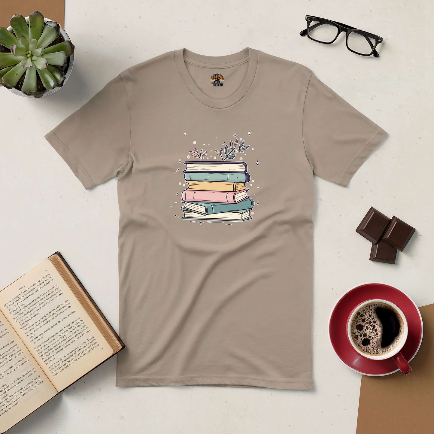 Blooming Books Tee with pastel books and floral design on a beige shirt, surrounded by coffee, chocolate, glasses, and an open book.