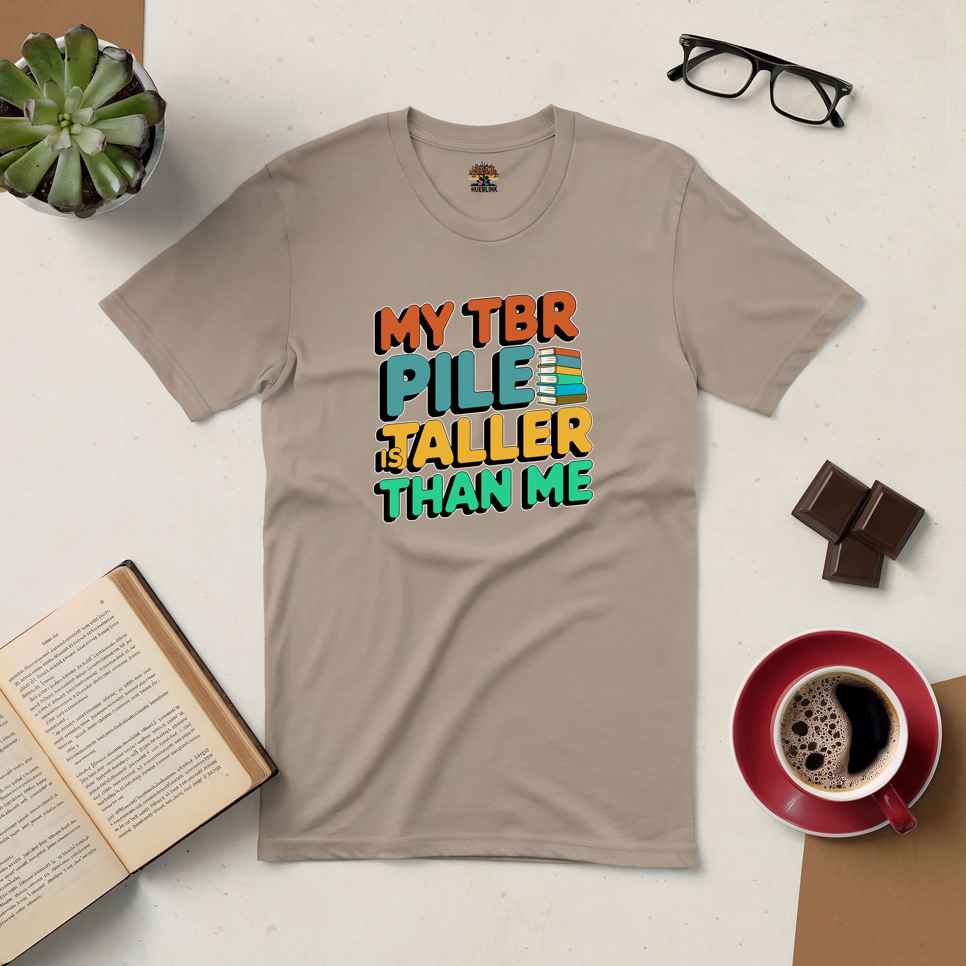 Beige t-shirt with "My TBR Pile is Taller Than Me" design surrounded by a book, coffee, chocolate, and glasses on a table.