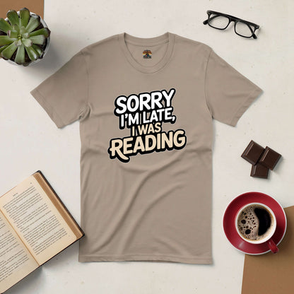 Beige t-shirt with "Sorry I'm Late, I Was Reading" text, surrounded by a book, glasses, coffee, and chocolate. Perfect for book lovers.