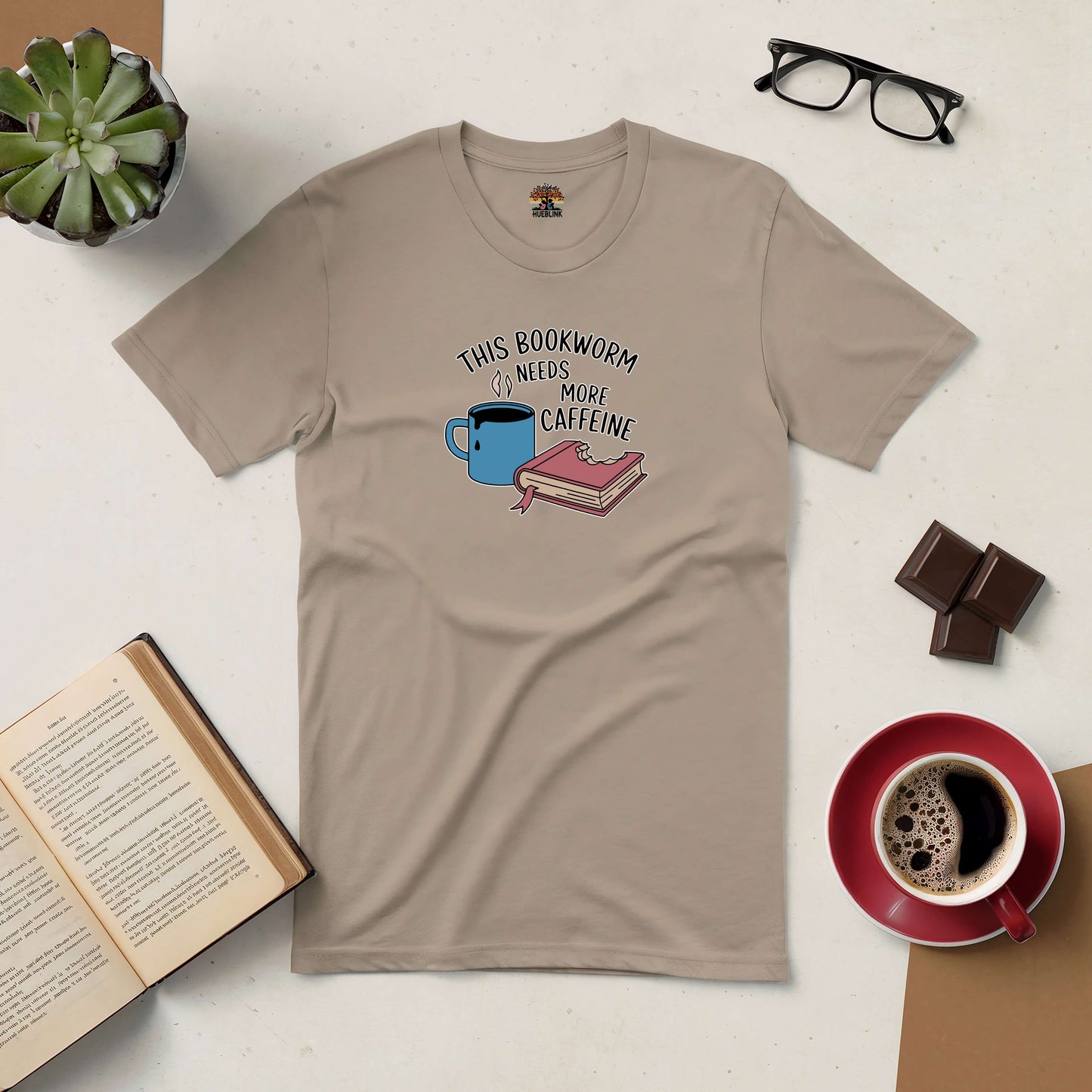 Beige t-shirt with "This Bookworm Needs More Caffeine" design, surrounded by a book, coffee cup, chocolate, glasses, and plant.