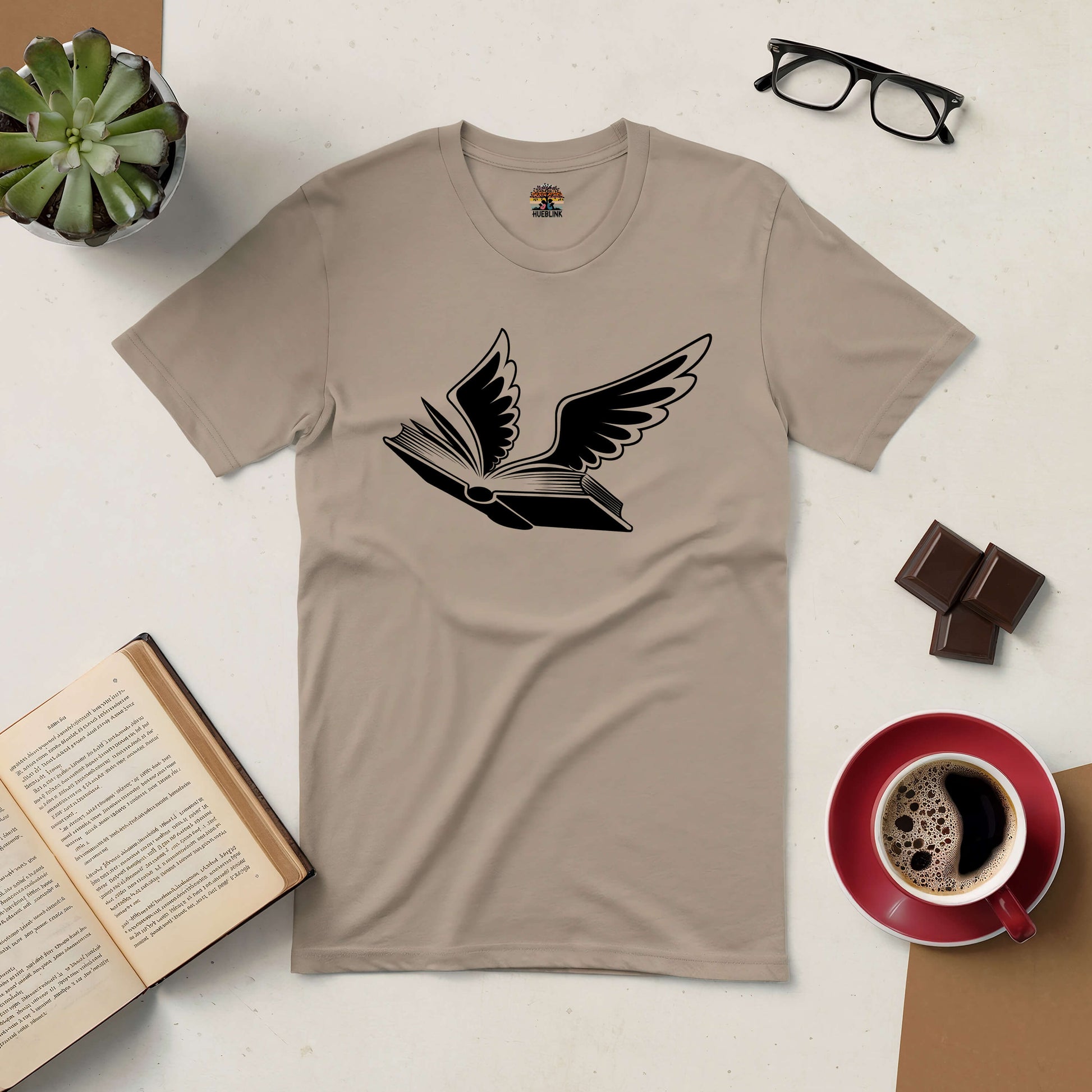 Bird of Hermes Tee with book and wings design, symbolizing soaring knowledge, surrounded by coffee, glasses, and an open book.