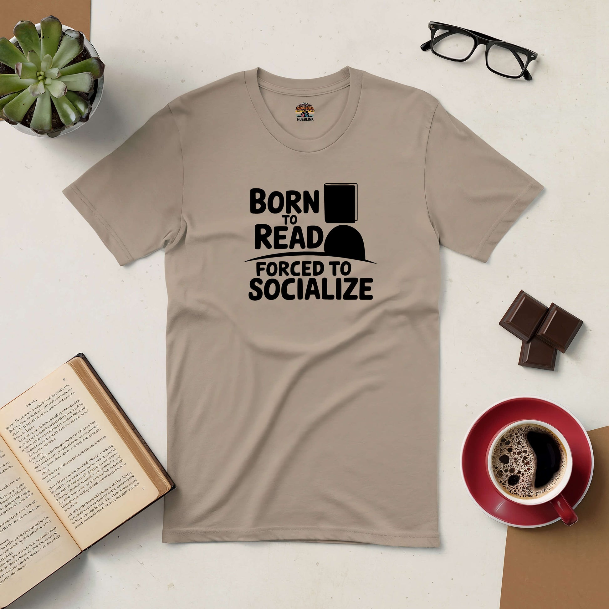 Brown "Born to Read, Forced to Socialize" tee for introverts displayed with glasses, open book, coffee, succulent, and chocolate.