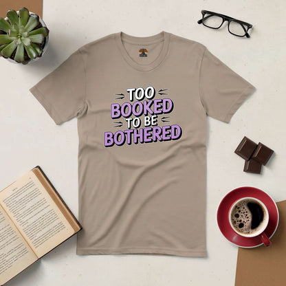 Beige "Too Booked to Be Bothered" tee beside a book, glasses, coffee, and chocolate, emphasizing reading and relaxation.