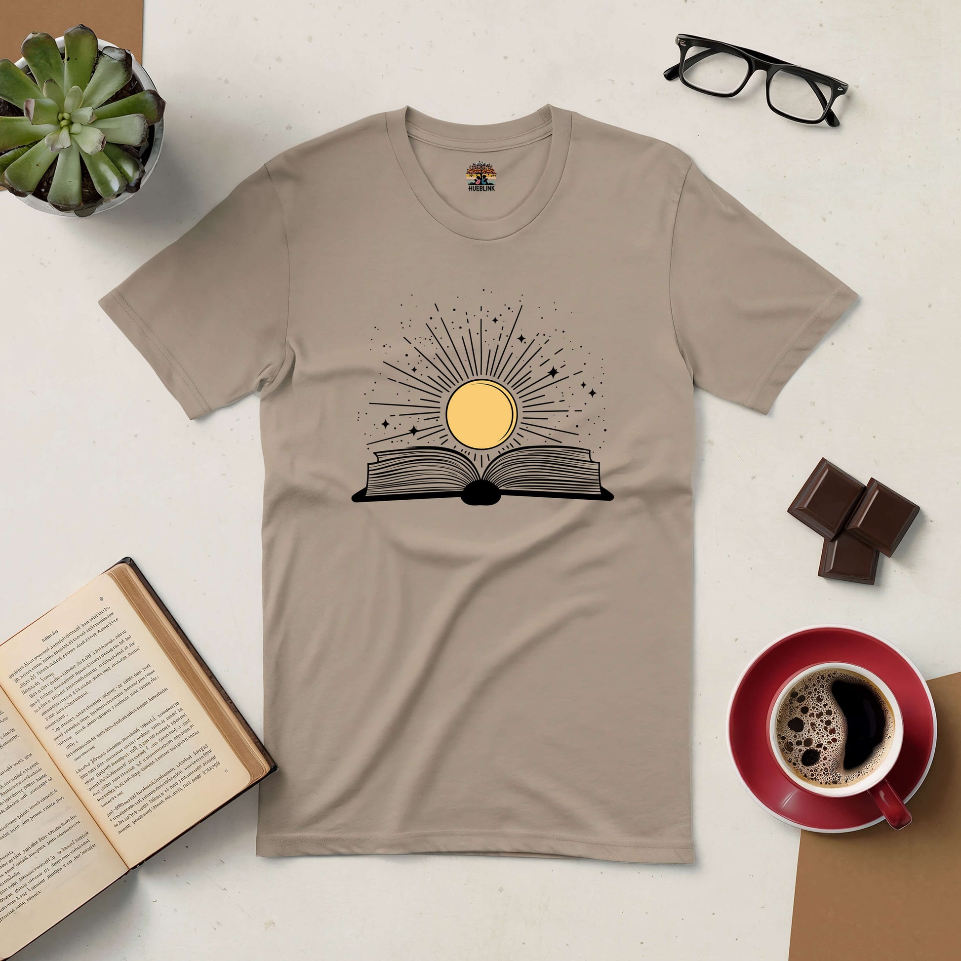 Sunshine Kiss Tee with book design, surrounded by coffee, chocolate, glasses, and plants on a light table.