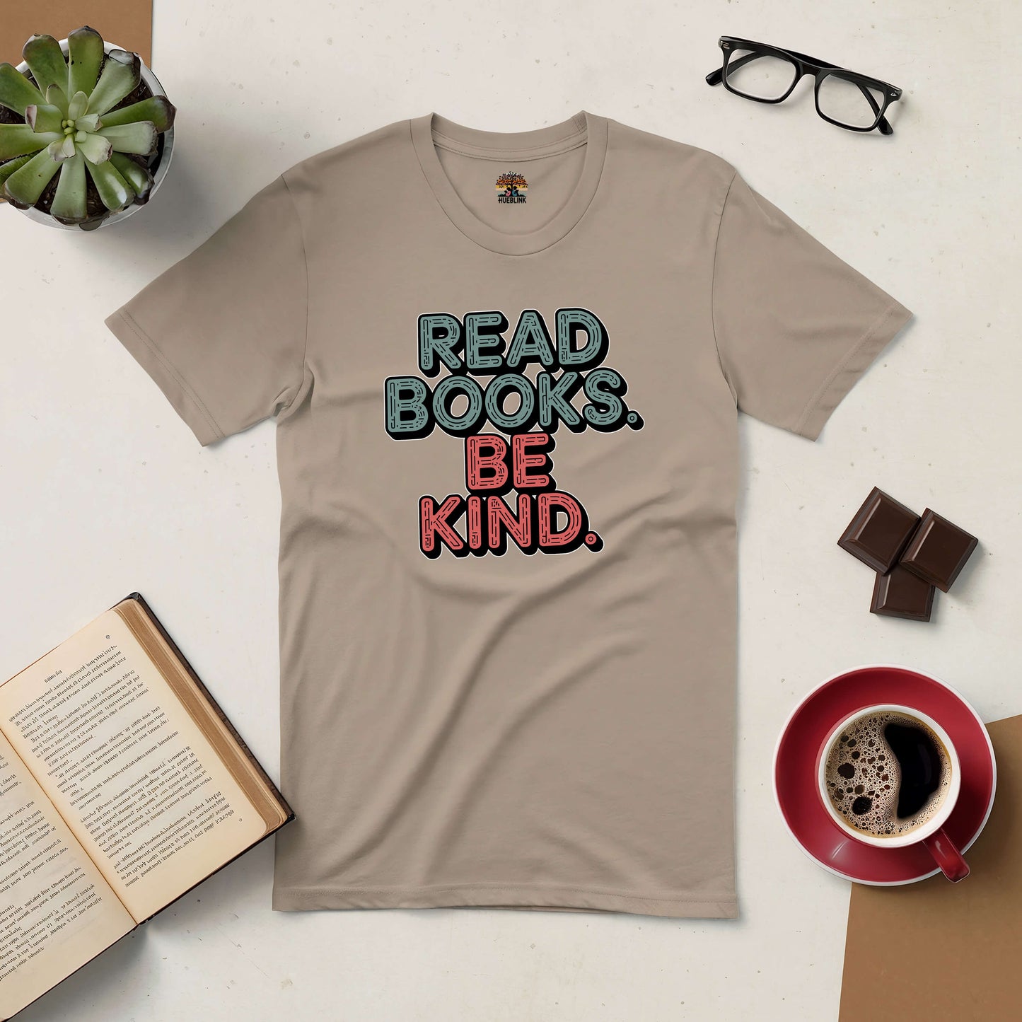 Beige "Read Books. Be Kind." tee for book lovers, surrounded by coffee, eyeglasses, chocolate, and an open book on a table.