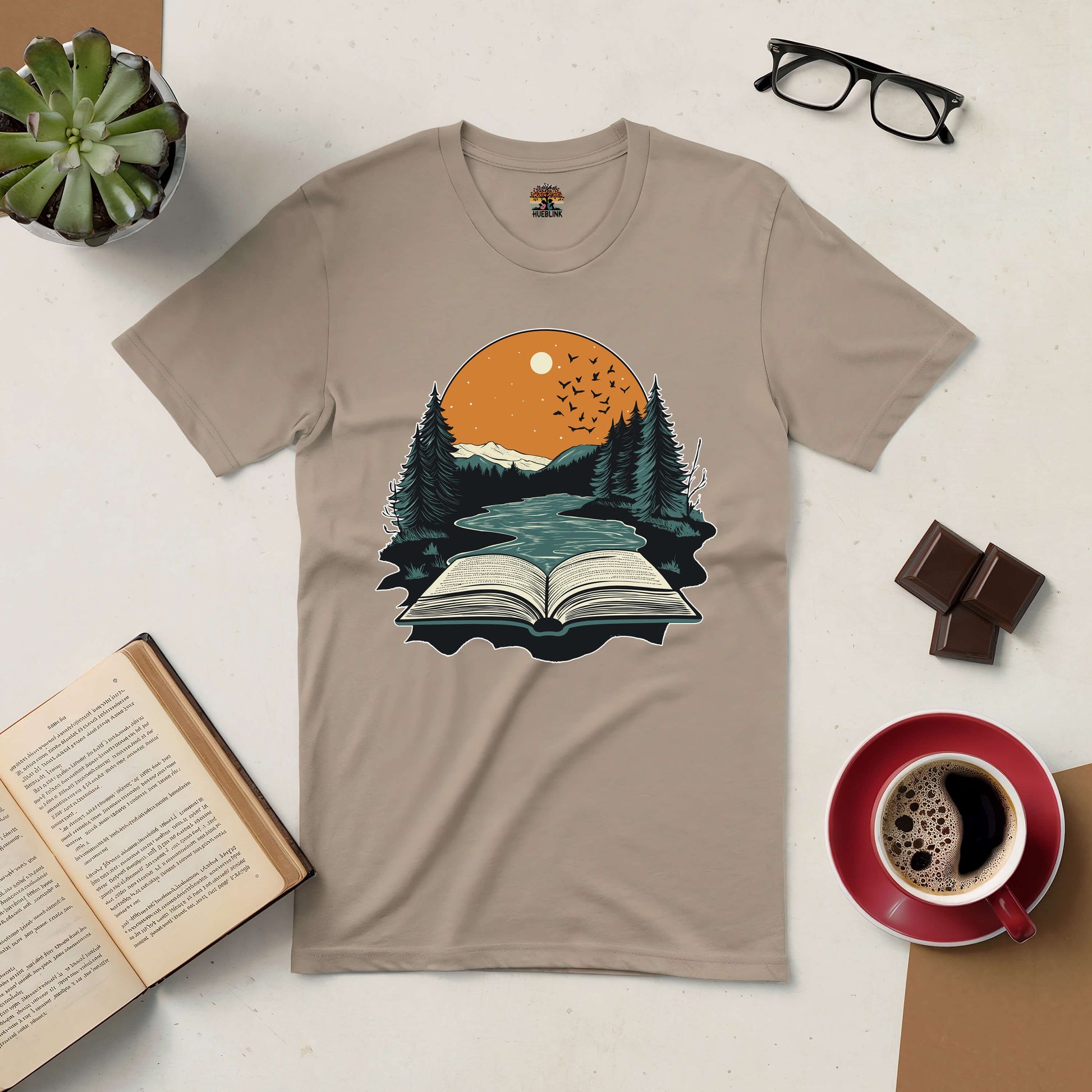 Beige "Absorbed in Nature" tee featuring an open book with forest and sunset illustration, surrounded by book, coffee, glasses, and plant.