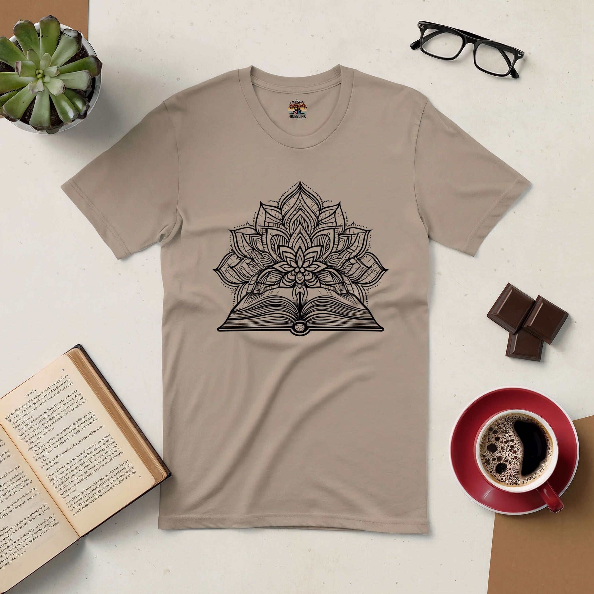 Beige t-shirt with intricate design of a blooming flower emerging from an open book, surrounded by coffee, chocolate, glasses, and a plant.