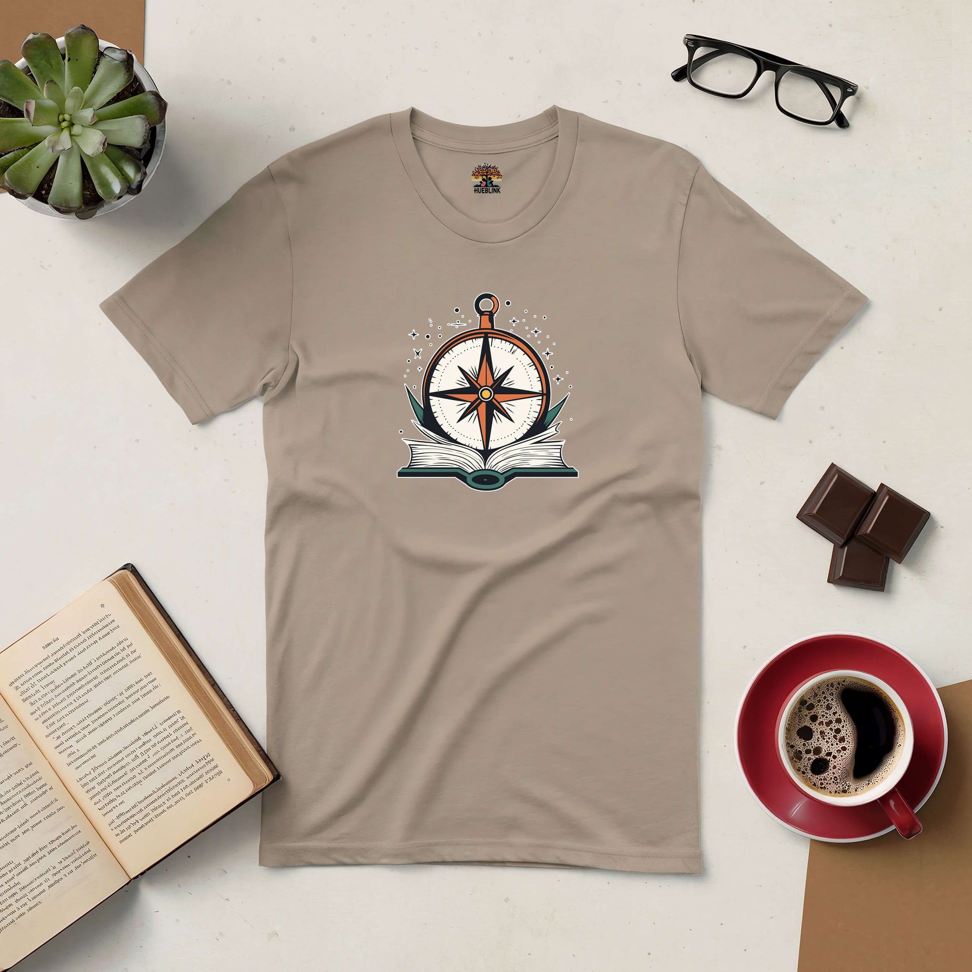 Beige T-shirt with compass and open book design. Coffee, glasses, and succulent in the background.