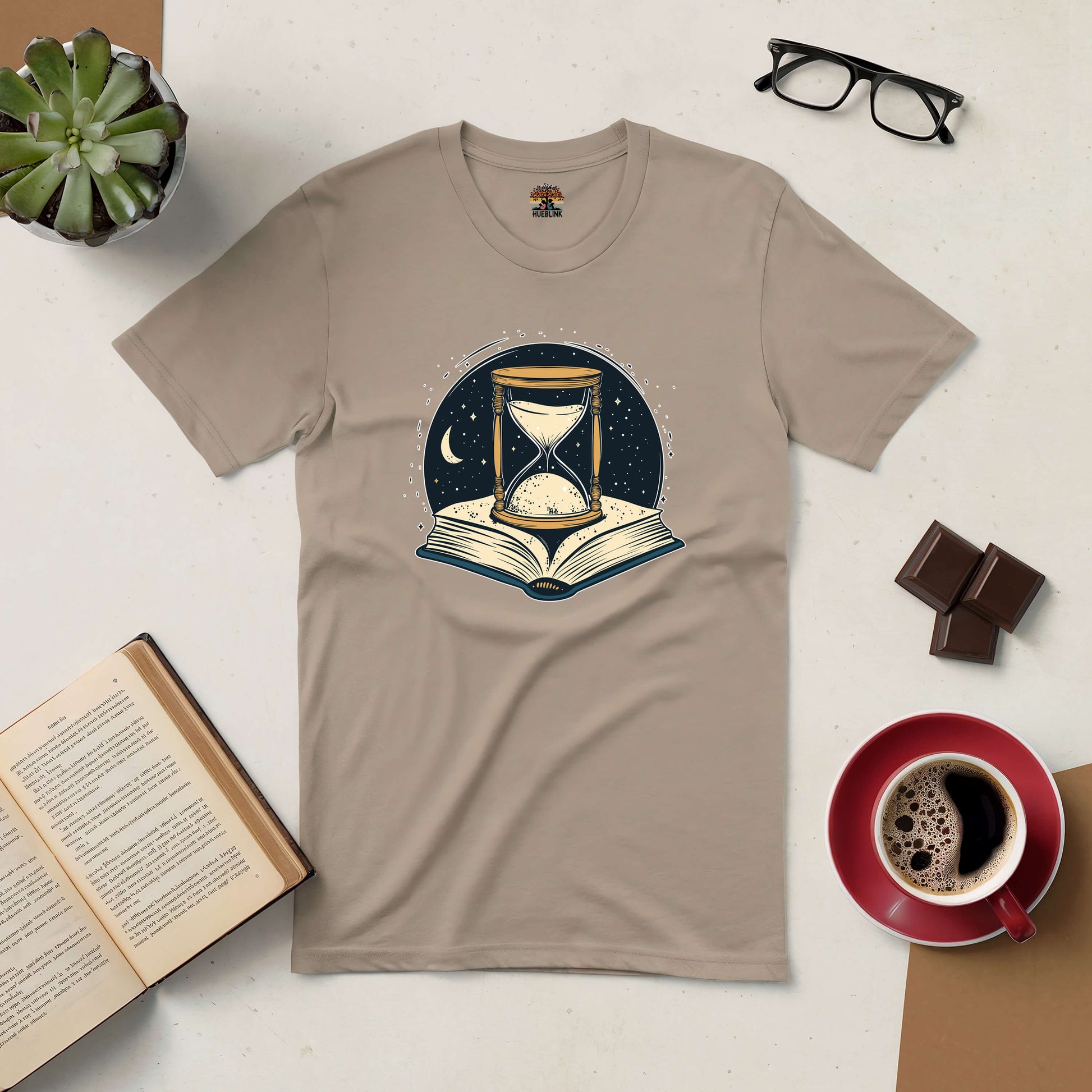 Beige t-shirt featuring an hourglass and open book design, symbolizing time and imagination, with coffee and book beside it.