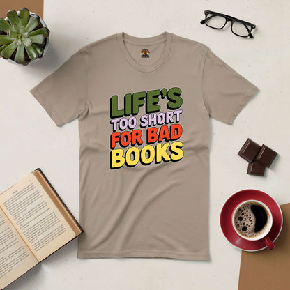 "Life's Too Short for Bad Books Tee with bold text on beige shirt, surrounded by coffee, glasses, chocolate, and an open book."