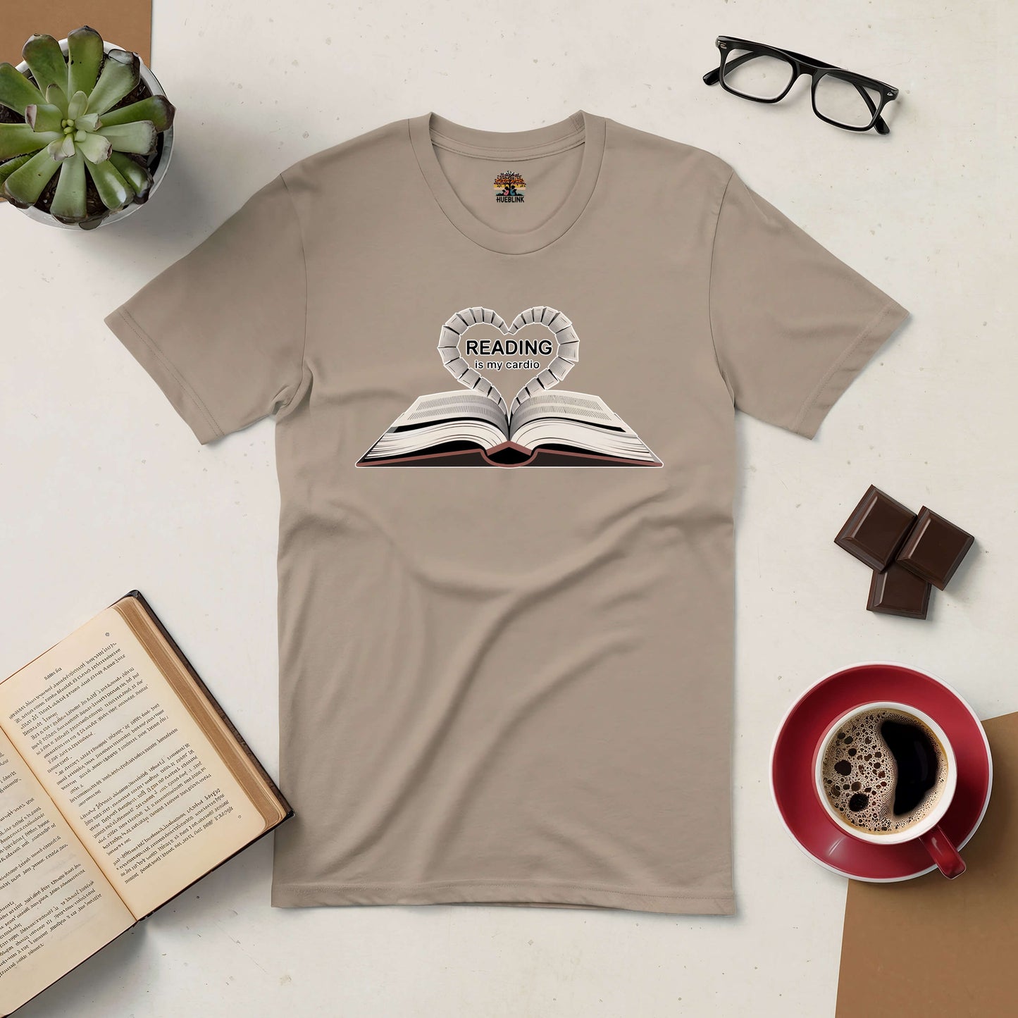 Beige "Reading Is My Cardio" tee with book graphic, surrounded by coffee, chocolate, open book, glasses, and plant. Perfect for book lovers.