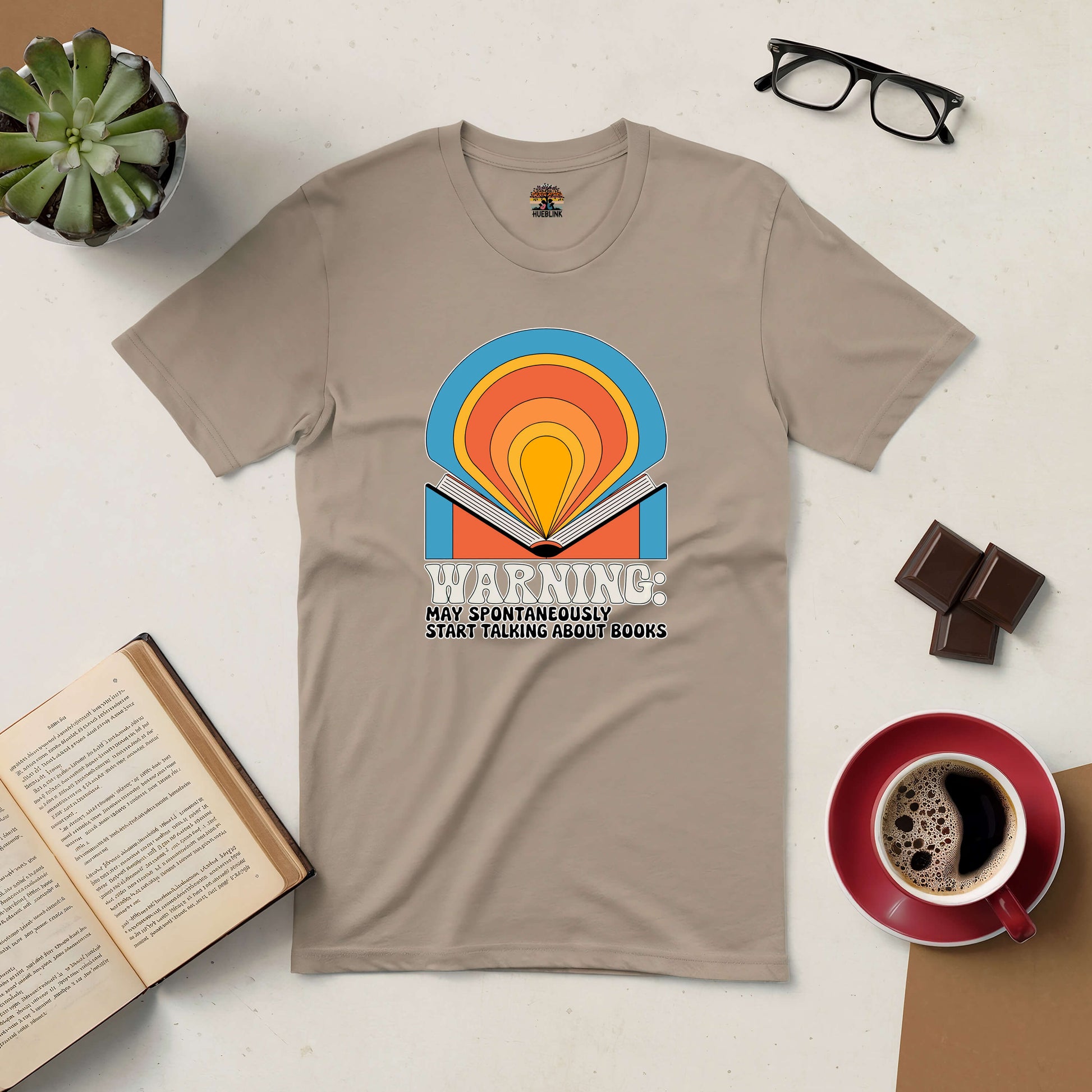 Beige tee with colorful book design and text "Warning: May Spontaneously Start Talking About Books," surrounded by coffee, glasses, and plant.