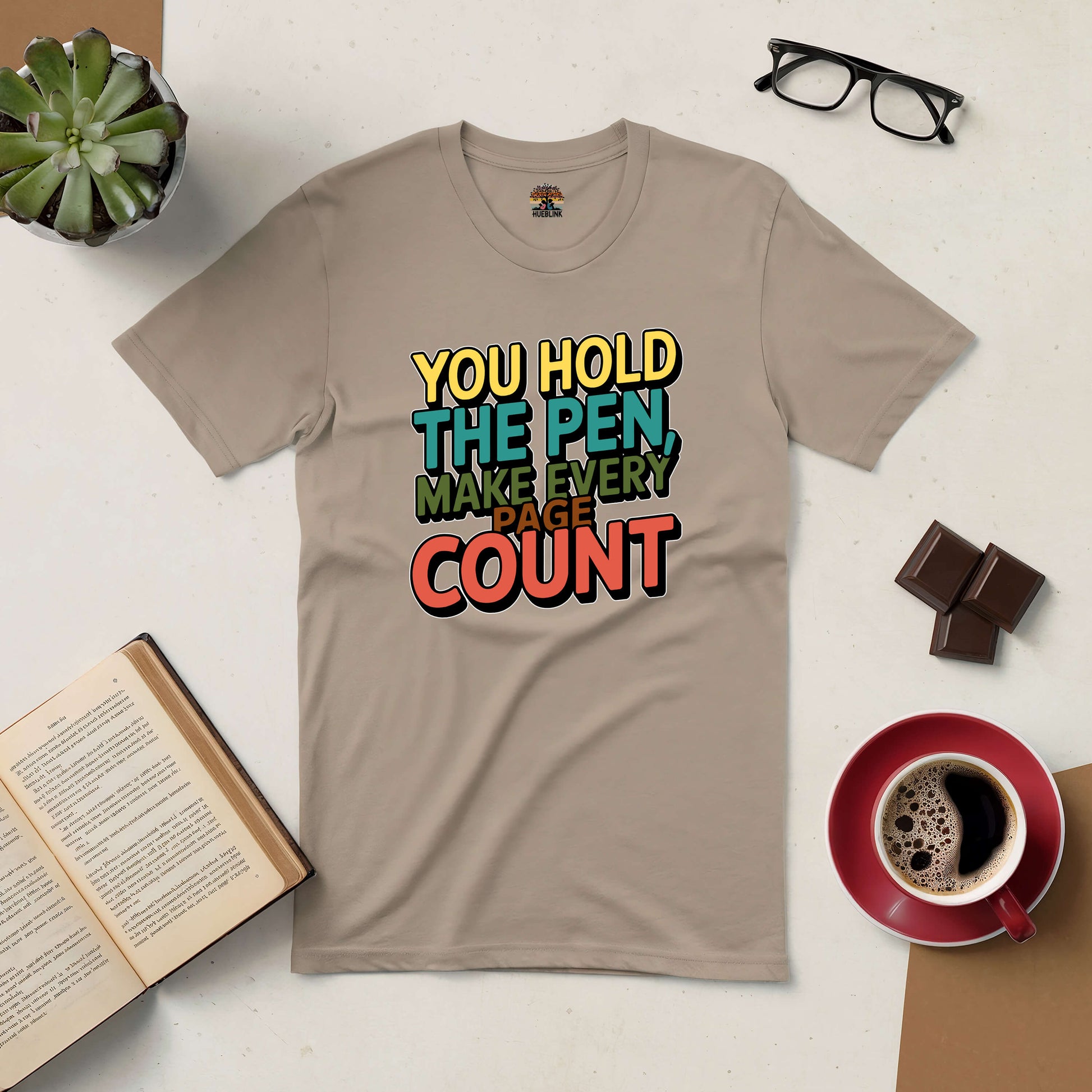 Inspirational tan t-shirt with text "You Hold the Pen, Make Every Page Count" surrounded by coffee, open book, plant, glasses, and chocolate.