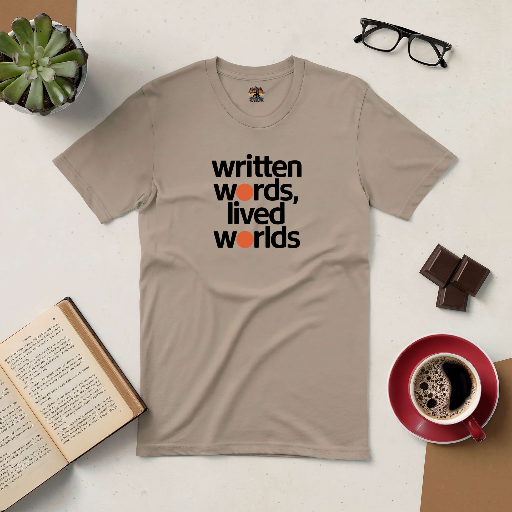 Beige tee with "Written Words, Lived Worlds" text, surrounded by an open book, coffee cup, chocolate, and eyeglasses on a table.