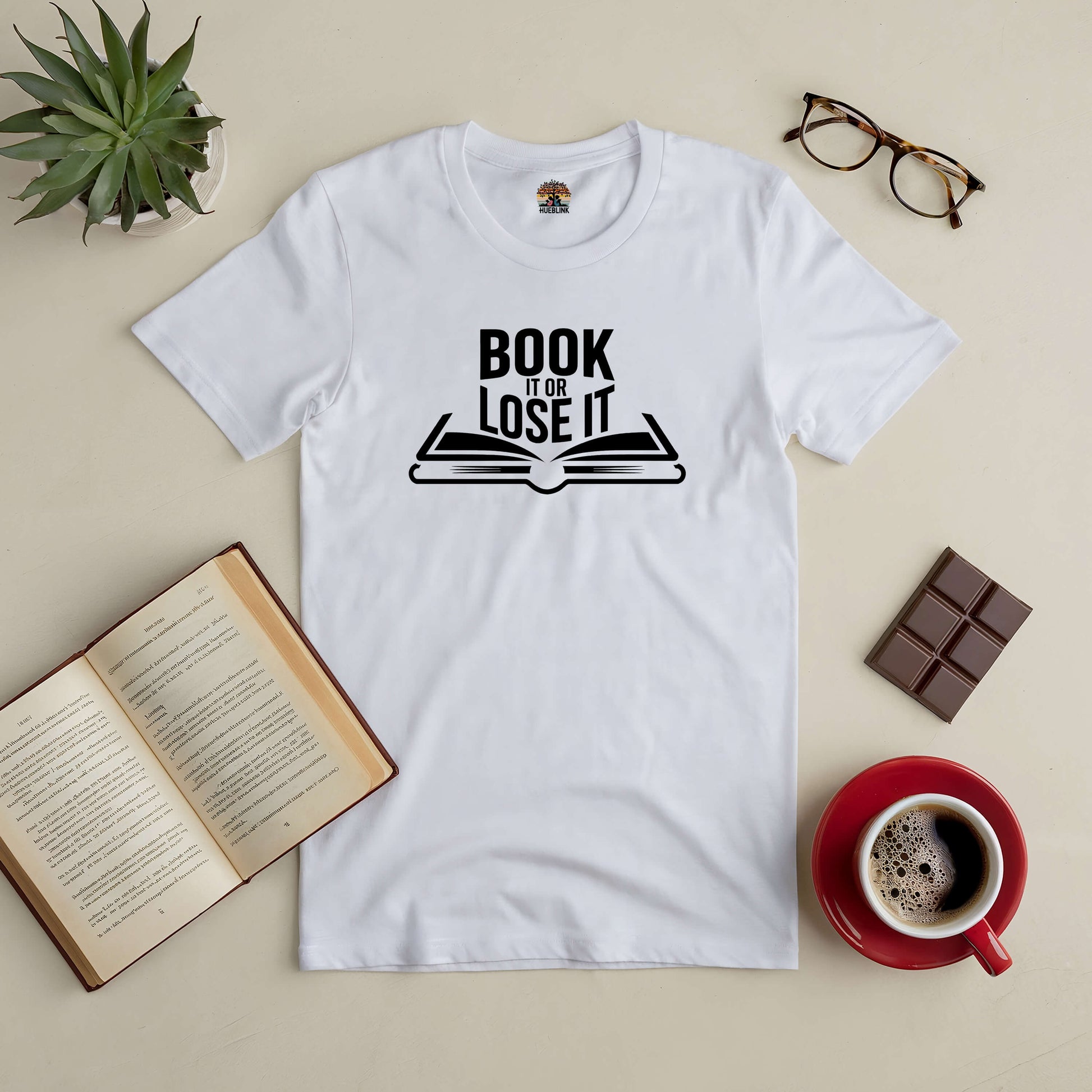 White "Book It or Lose It" tee with open book design, surrounded by an open book, glasses, chocolate, and a coffee cup.