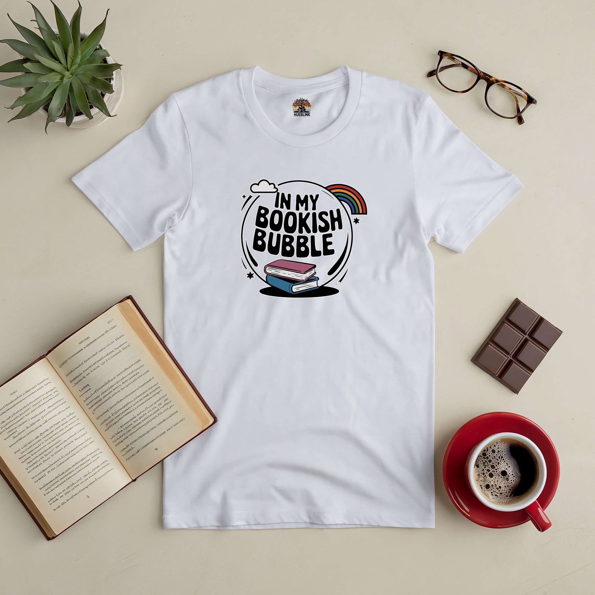 White "In My Bookish Bubble" Tee surrounded by open book, coffee, chocolate, glasses, and plant on a beige background.