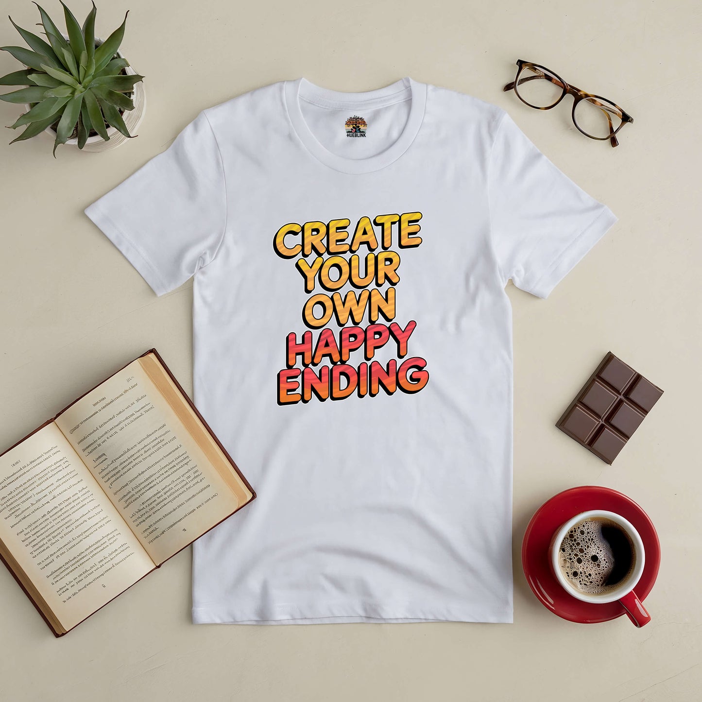 White tee with "Create Your Own Happy Ending" text, surrounded by glasses, plant, book, chocolate, and coffee cup on a beige background.