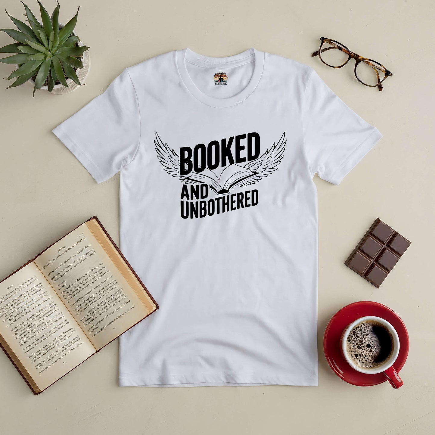 White "Booked and Unbothered" tee with open book and wings design, surrounded by a book, glasses, coffee, and chocolate.