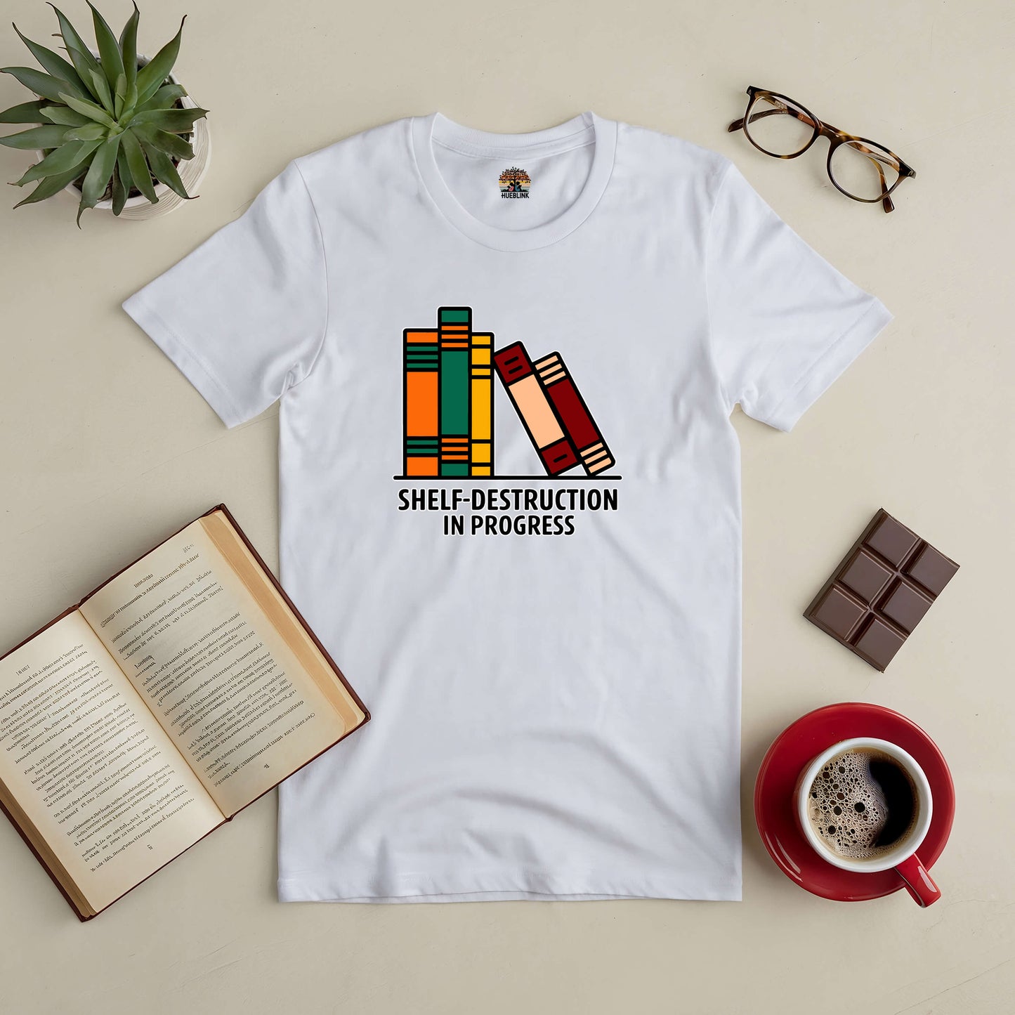 White "Shelf-Destruction in Progress" tee with book design, surrounded by open book, coffee cup, glasses, chocolate, and plant.