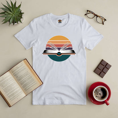 Gateway to New Horizons Tee with book graphic, surrounded by open book, coffee, glasses, plant, and chocolate on a beige table.