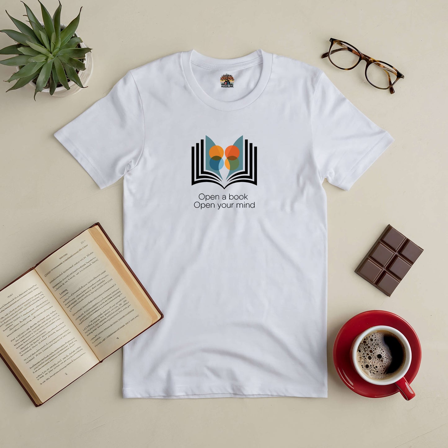 White "Open a Book, Open Your Mind" tee with book design, surrounded by an open book, coffee, chocolate, glasses, and a plant.