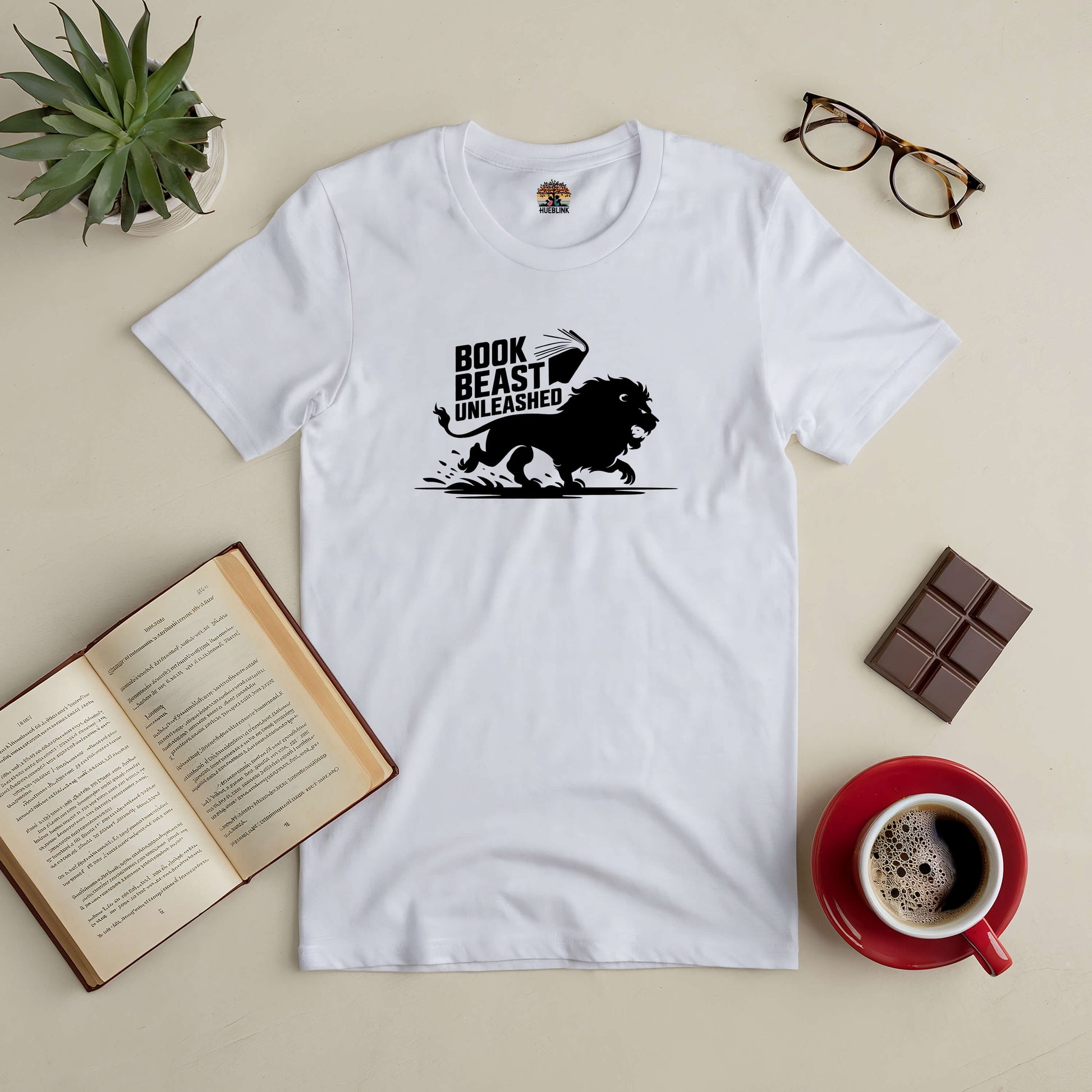 White "Book Beast Unleashed" tee with roaring lion design, placed with an open book, coffee, glasses, and chocolate bar.