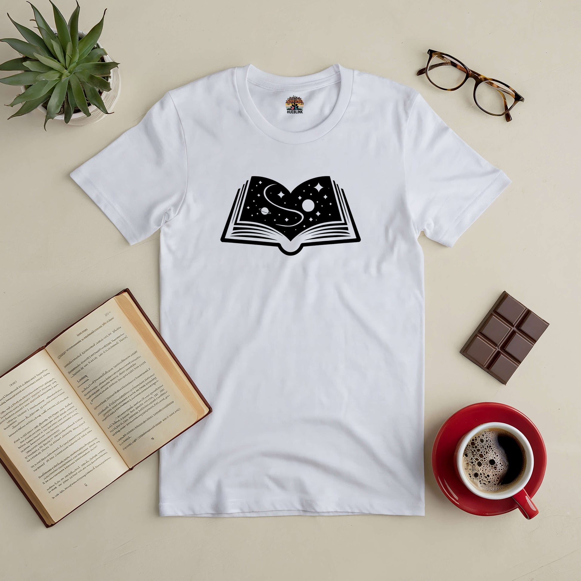 "Books: A Universe Within Tee featuring a cosmic book design, surrounded by a book, glasses, chocolate, and a coffee on a table."